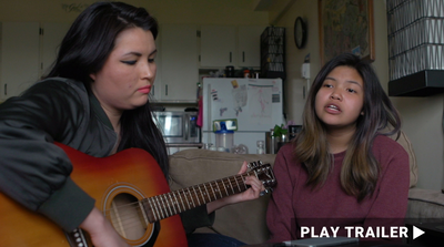 Trailer for documentary "Another Word For Learning" directed by Jadis Dumas. Woman playing orange guitar with girl singing on couch. https://vimeo.com/694247624