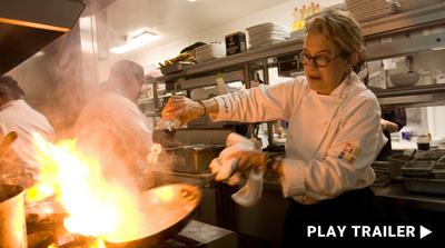 Trailer for documentary “SUSAN FENIGER. FORKED” directed by Liz Lachman. A female cook is holding a pan and pouring sauce, a bright fire on the stove. https://vimeo.com/1012585875