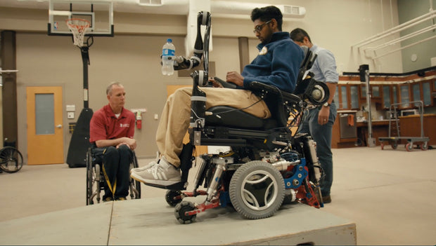Still from the documentary "Bumps in the Road." Person using a wheelchair with a mechanical arm in an indoor setting
